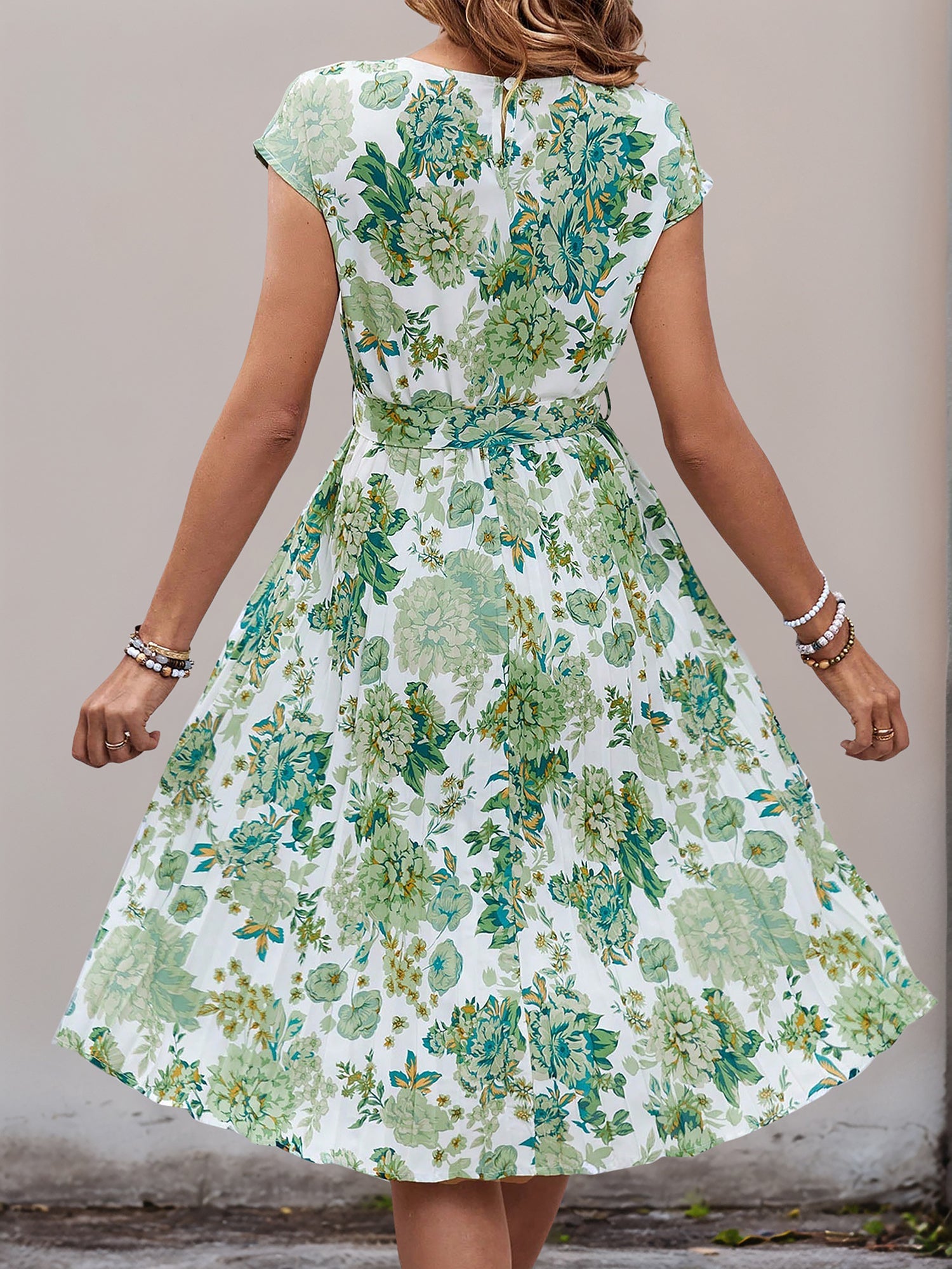 Green - Woman wearing a lightweight floral cap sleeve midi dress with a tie waist and pleated skirt.