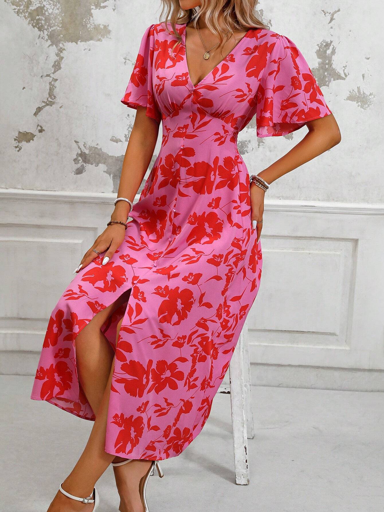 Pink - A woman wearing a pink and red floral print maxi dress with flutter sleeves, a V-neckline, button-front detailing, and a high front slit, holding a woven straw handbag.