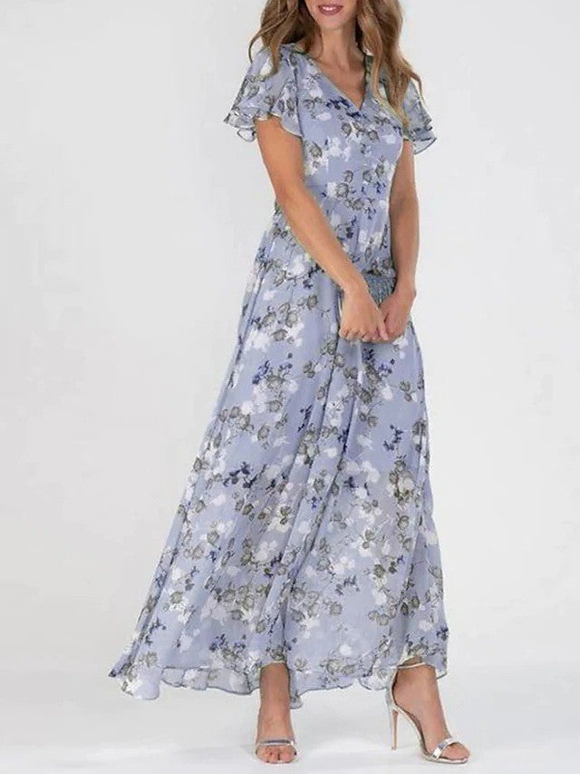 Lilac - A woman wearing a dusty blue floral print chiffon maxi dress with flutter sleeves, a V-neckline, and a flowing skirt, paired with silver heeled sandals.