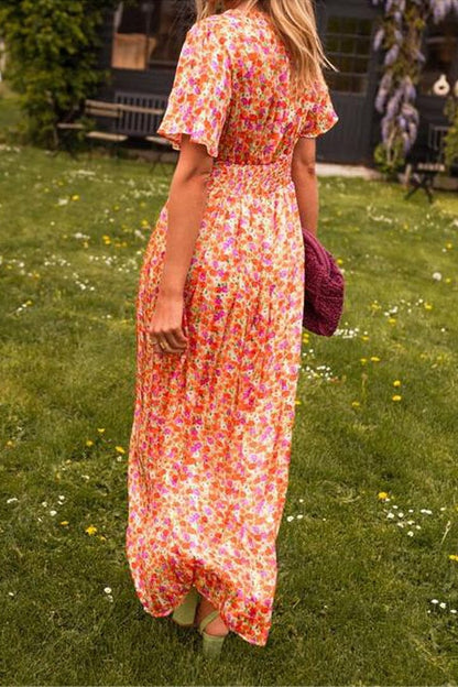 Orange - A woman wearing a vibrant orange and pink floral print maxi dress with flutter sleeves, a plunging V-neckline, and a ruched waistband, standing on a green lawn.
