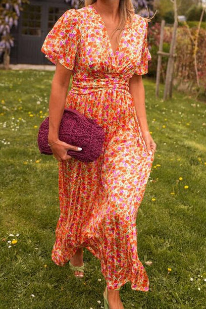 Orange - A woman wearing a vibrant orange and pink floral print maxi dress with flutter sleeves, a plunging V-neckline, and a ruched waistband, standing on a green lawn.