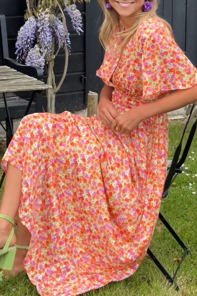 Orange - A woman wearing a vibrant orange and pink floral print maxi dress with flutter sleeves, a plunging V-neckline, and a ruched waistband, standing on a green lawn.