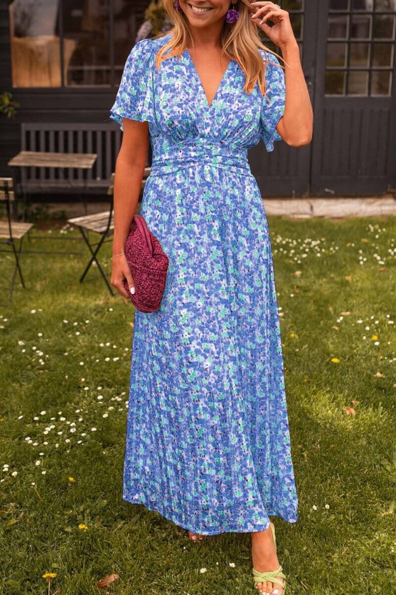 Blue - A woman wearing a vibrant orange and pink floral print maxi dress with flutter sleeves, a plunging V-neckline, and a ruched waistband, standing on a green lawn.