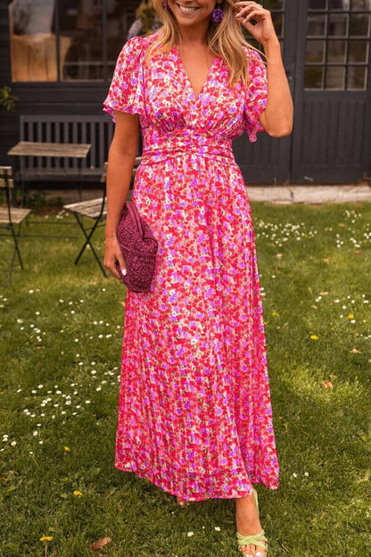 Pink - A woman wearing a vibrant orange and pink floral print maxi dress with flutter sleeves, a plunging V-neckline, and a ruched waistband, standing on a green lawn.