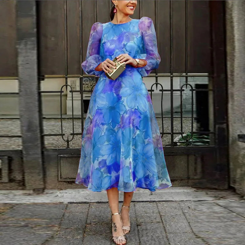 Blue - Woman wearing a vibrant pink floral chiffon midi dress with sheer puff sleeves and an A-line skirt, walking outdoors holding a gold clutch.
