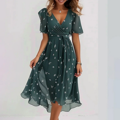 Green - A woman wearing a dark green chiffon midi dress with a white ditsy floral print, featuring flutter sleeves, a surplice V-neckline, a self-tie waist, and a flowing curved hem.