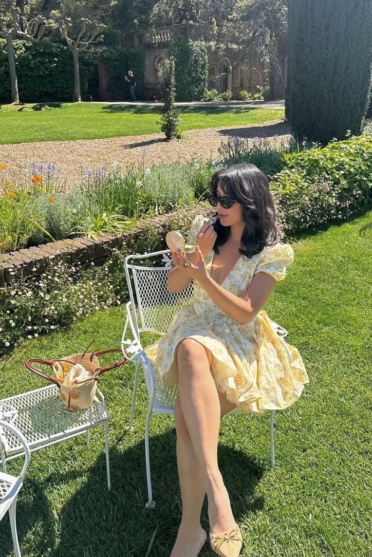 Yellow - Woman wearing a yellow ditsy floral mini dress featuring a plunging neckline, tie-front corset bodice, short puff sleeves, and a flared gathered skirt, standing outdoors on a sunny patio.