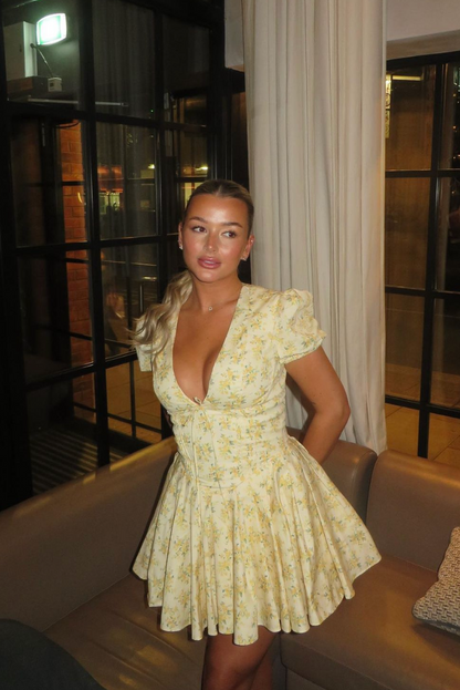 Yellow - Woman wearing a yellow ditsy floral mini dress featuring a plunging neckline, tie-front corset bodice, short puff sleeves, and a flared gathered skirt, standing outdoors on a sunny patio.