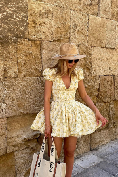 Yellow - Woman wearing a yellow ditsy floral mini dress featuring a plunging neckline, tie-front corset bodice, short puff sleeves, and a flared gathered skirt, standing outdoors on a sunny patio.
