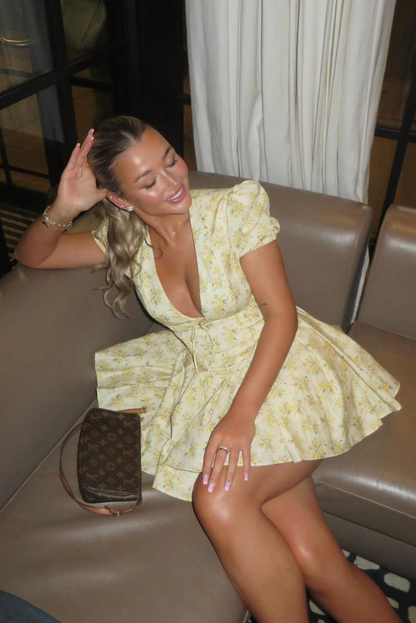 Yellow - Woman wearing a yellow ditsy floral mini dress featuring a plunging neckline, tie-front corset bodice, short puff sleeves, and a flared gathered skirt, standing outdoors on a sunny patio.