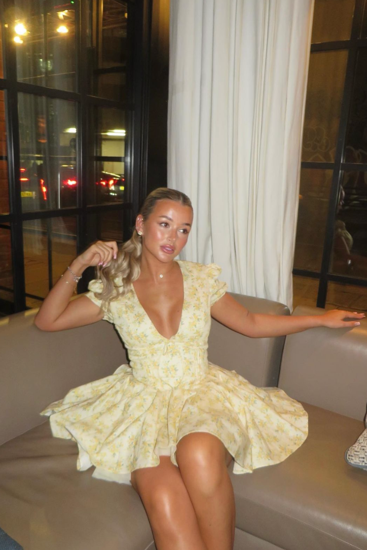 Yellow - Woman wearing a yellow ditsy floral mini dress featuring a plunging neckline, tie-front corset bodice, short puff sleeves, and a flared gathered skirt, standing outdoors on a sunny patio.