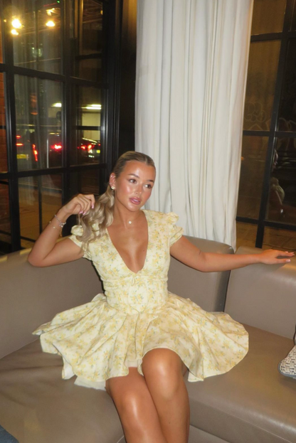 Yellow - Woman wearing a yellow ditsy floral mini dress featuring a plunging neckline, tie-front corset bodice, short puff sleeves, and a flared gathered skirt, standing outdoors on a sunny patio.