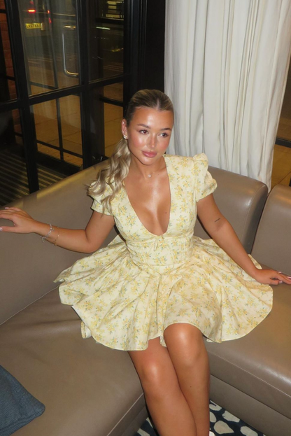 Yellow - Woman wearing a yellow ditsy floral mini dress featuring a plunging neckline, tie-front corset bodice, short puff sleeves, and a flared gathered skirt, standing outdoors on a sunny patio.