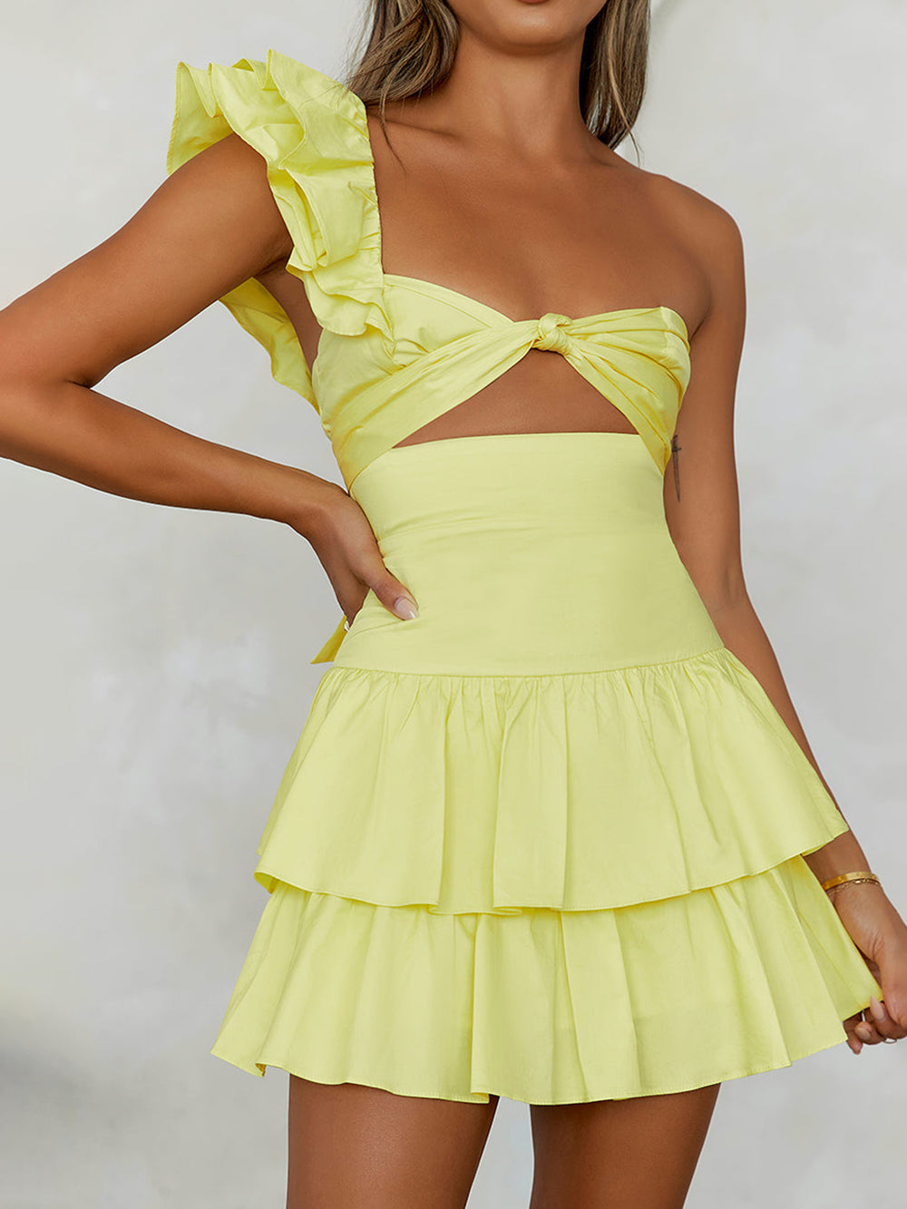 A woman wearing a pink and white ditsy floral print mini dress featuring a one-shoulder ruffled strap, a front cutout at the waist, and a tiered ruffled skirt.