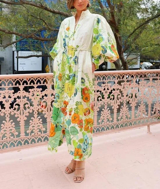 A woman wearing a cream maxi dress featuring a vibrant botanical floral print in green, yellow, and orange, designed with voluminous balloon sleeves, a stand collar, and a thin waist tie.