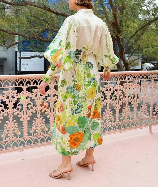 A woman wearing a cream maxi dress featuring a vibrant botanical floral print in green, yellow, and orange, designed with voluminous balloon sleeves, a stand collar, and a thin waist tie.