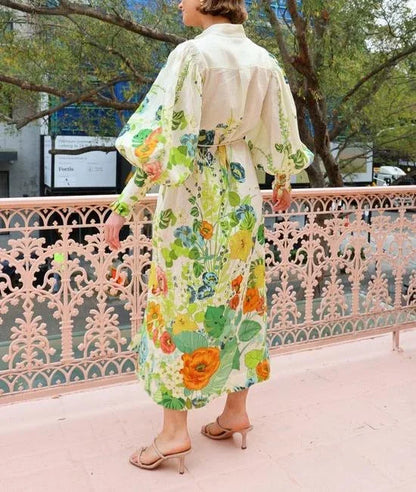 A woman wearing a cream maxi dress featuring a vibrant botanical floral print in green, yellow, and orange, designed with voluminous balloon sleeves, a stand collar, and a thin waist tie.