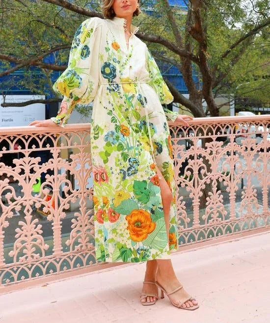 A woman wearing a cream maxi dress featuring a vibrant botanical floral print in green, yellow, and orange, designed with voluminous balloon sleeves, a stand collar, and a thin waist tie.