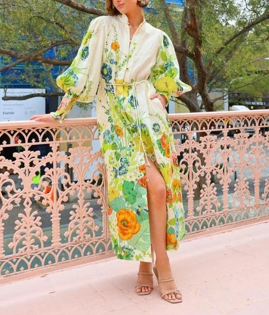 A woman wearing a cream maxi dress featuring a vibrant botanical floral print in green, yellow, and orange, designed with voluminous balloon sleeves, a stand collar, and a thin waist tie.