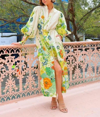 A woman wearing a cream maxi dress featuring a vibrant botanical floral print in green, yellow, and orange, designed with voluminous balloon sleeves, a stand collar, and a thin waist tie.