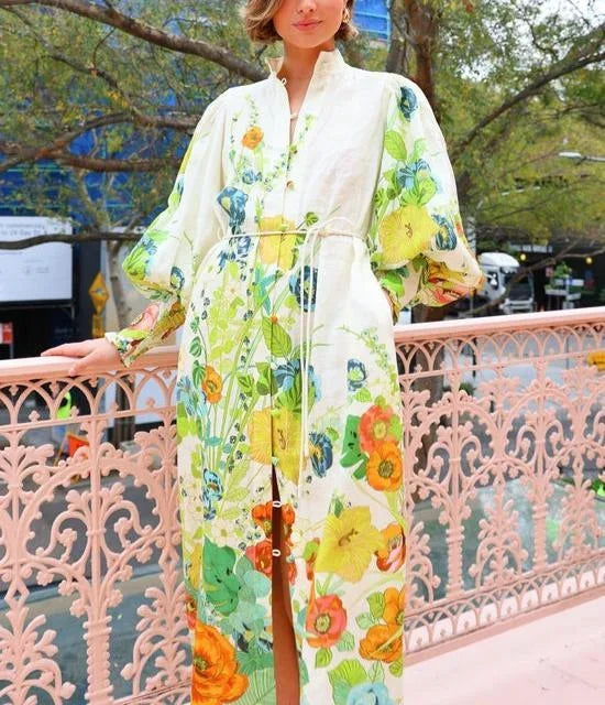 Flowery - A woman wearing a cream maxi dress featuring a vibrant botanical floral print in green, yellow, and orange, designed with voluminous balloon sleeves, a stand collar, and a thin waist tie.