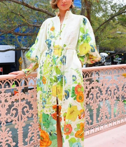 Flowery - A woman wearing a cream maxi dress featuring a vibrant botanical floral print in green, yellow, and orange, designed with voluminous balloon sleeves, a stand collar, and a thin waist tie.
