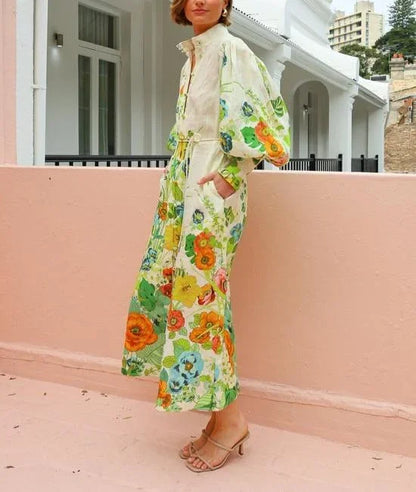 Flowery - A woman wearing a cream maxi dress featuring a vibrant botanical floral print in green, yellow, and orange, designed with voluminous balloon sleeves, a stand collar, and a thin waist tie.