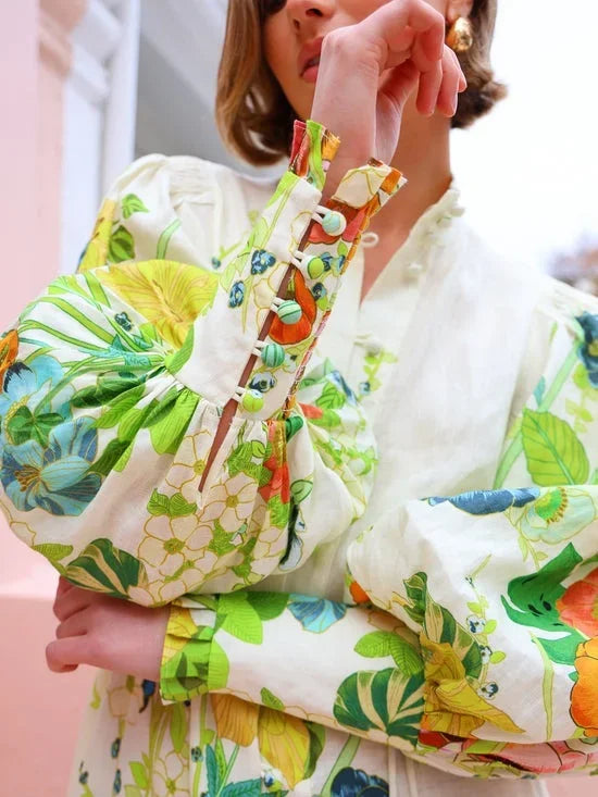 Flowery - A woman wearing a cream maxi dress featuring a vibrant botanical floral print in green, yellow, and orange, designed with voluminous balloon sleeves, a stand collar, and a thin waist tie.