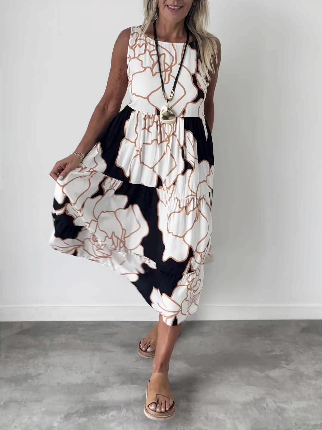 White - A woman wearing a sleeveless, tiered midi dress featuring a large-scale black and white floral outline print with tan accents, styled with a gold pendant necklace.