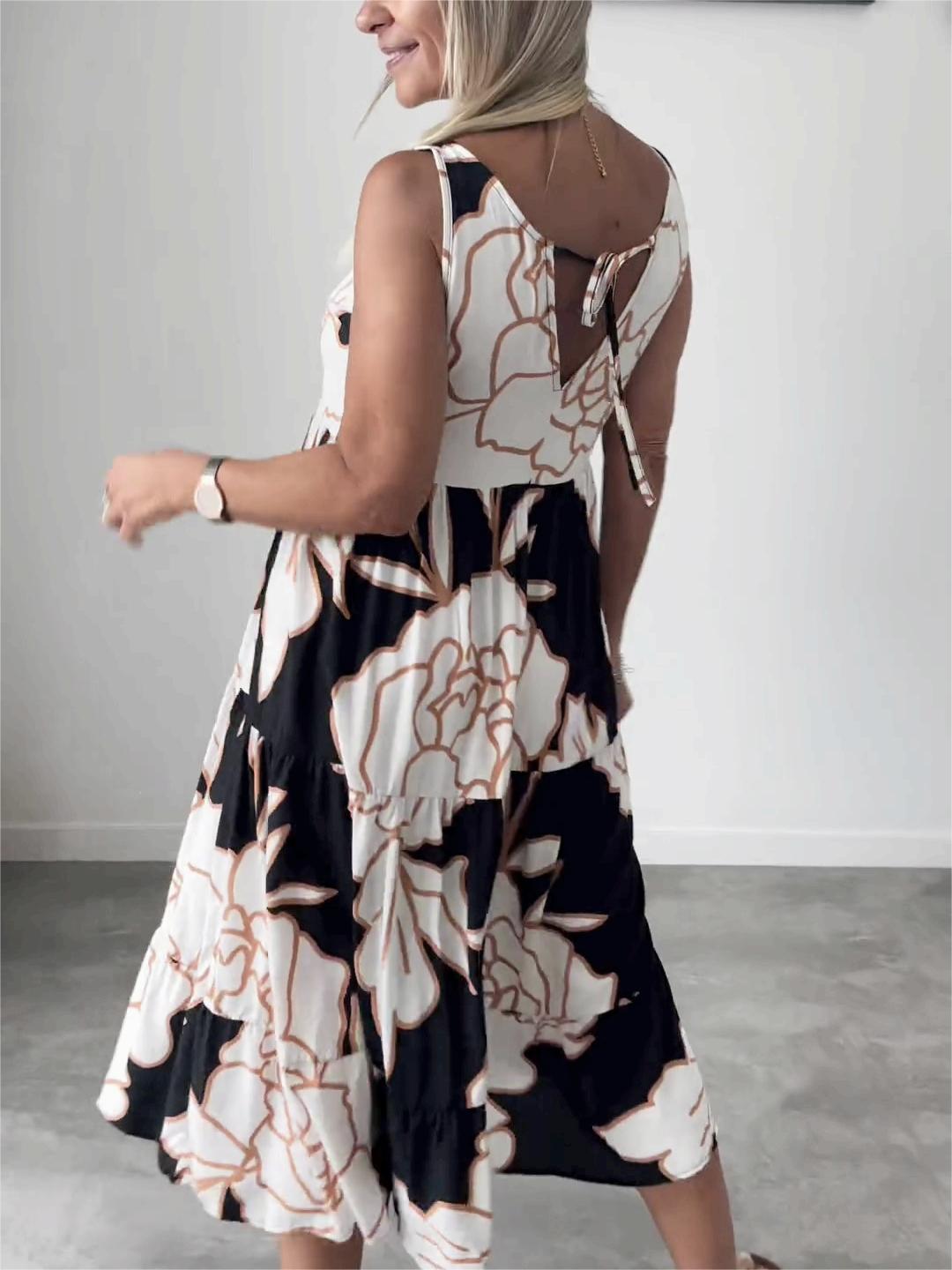 White - A woman wearing a sleeveless, tiered midi dress featuring a large-scale black and white floral outline print with tan accents, styled with a gold pendant necklace.