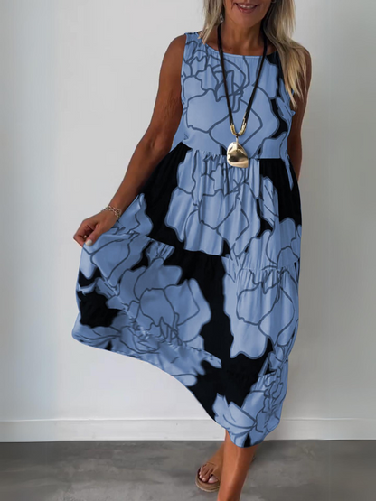 Blue - A woman wearing a sleeveless, tiered midi dress featuring a large-scale black and white floral outline print with tan accents, styled with a gold pendant necklace.