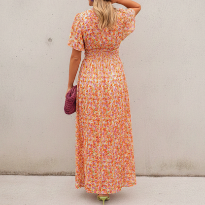 Orange - A woman wearing a vibrant orange and pink floral print maxi dress featuring a V-neckline, flutter sleeves, and a ruched waistband, standing against a neutral concrete wall.