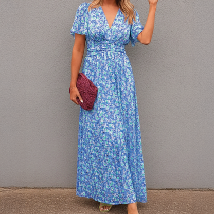 Blue - A woman wearing a vibrant orange and pink floral print maxi dress featuring a V-neckline, flutter sleeves, and a ruched waistband, standing against a neutral concrete wall.