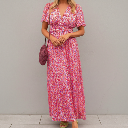 Pink - A woman wearing a vibrant orange and pink floral print maxi dress featuring a V-neckline, flutter sleeves, and a ruched waistband, standing against a neutral concrete wall.