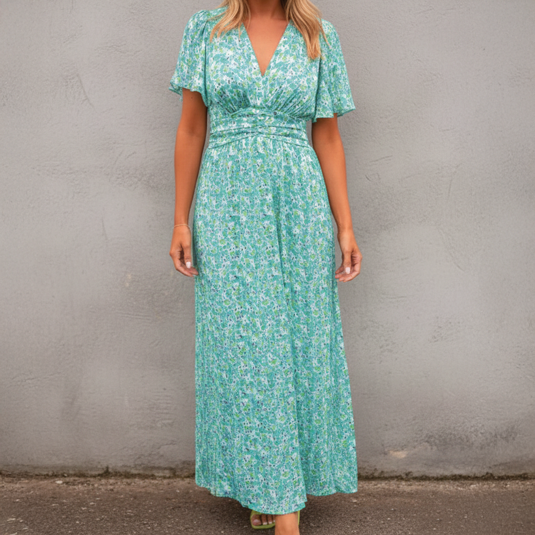 Green - A woman wearing a vibrant orange and pink floral print maxi dress featuring a V-neckline, flutter sleeves, and a ruched waistband, standing against a neutral concrete wall.