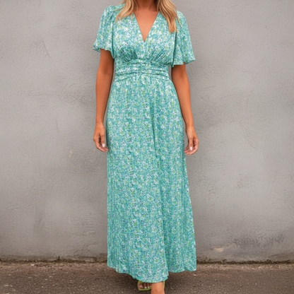 Green - A woman wearing a vibrant orange and pink floral print maxi dress featuring a V-neckline, flutter sleeves, and a ruched waistband, standing against a neutral concrete wall.