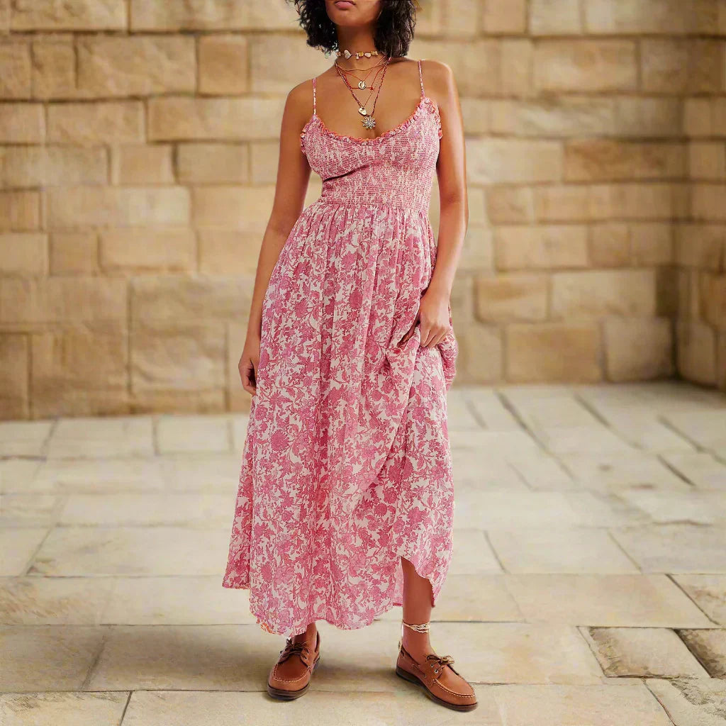 Floral Pink - A woman wearing a pink and white floral print maxi dress featuring a smocked bodice, ruffled neckline, and thin straps, standing against a rustic stone wall.