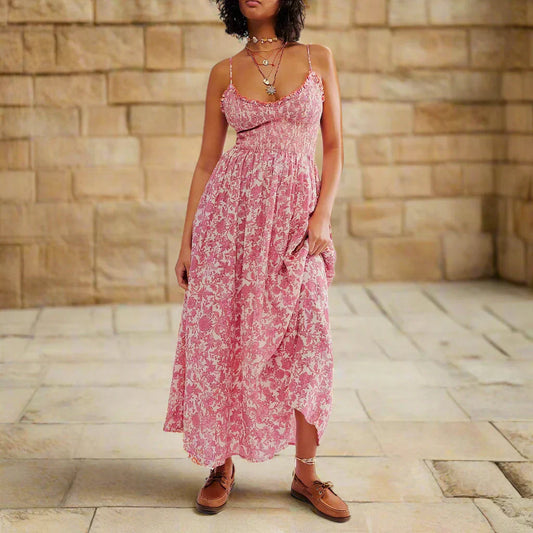 Floral Pink - A woman wearing a pink and white floral print maxi dress featuring a smocked bodice, ruffled neckline, and thin straps, standing against a rustic stone wall.