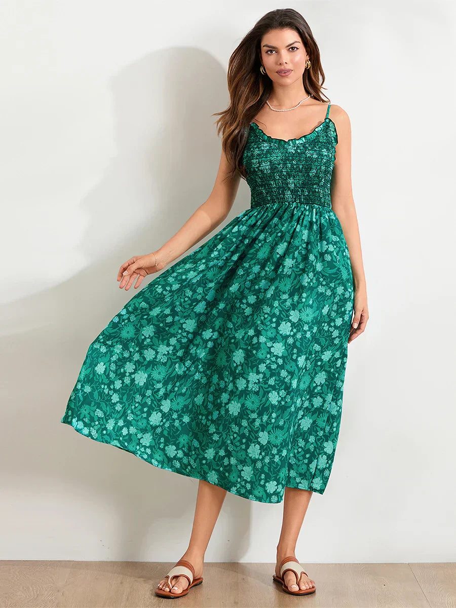 Floral Cyan - A woman wearing a pink and white floral print maxi dress featuring a smocked bodice, ruffled neckline, and thin straps, standing against a rustic stone wall.