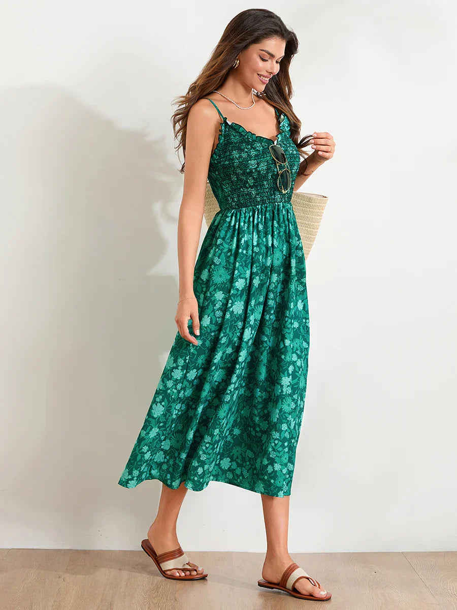 Floral Cyan - A woman wearing a pink and white floral print maxi dress featuring a smocked bodice, ruffled neckline, and thin straps, standing against a rustic stone wall.