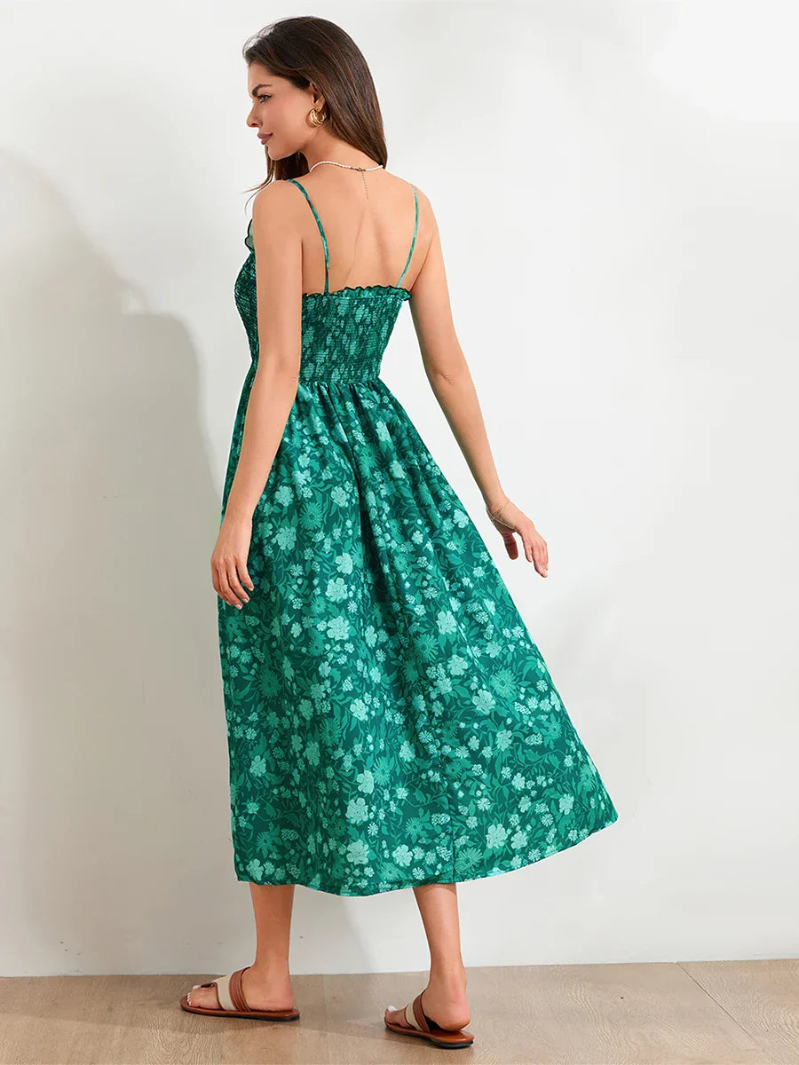 Floral Cyan - A woman wearing a pink and white floral print maxi dress featuring a smocked bodice, ruffled neckline, and thin straps, standing against a rustic stone wall.