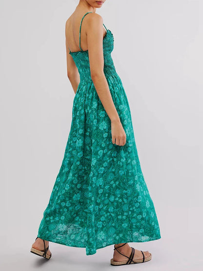 Floral Cyan - A woman wearing a pink and white floral print maxi dress featuring a smocked bodice, ruffled neckline, and thin straps, standing against a rustic stone wall.
