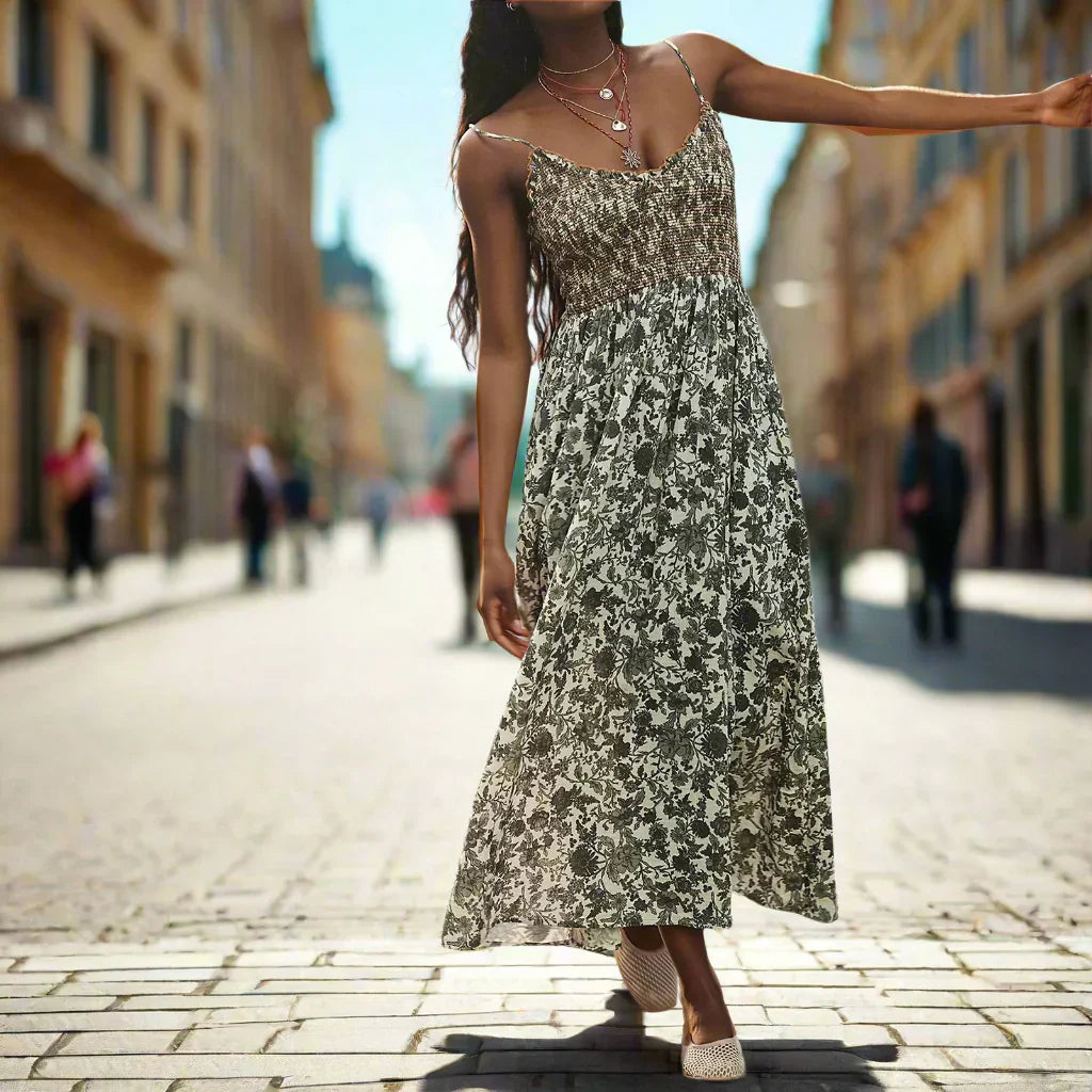 Floral Black - A woman wearing a pink and white floral print maxi dress featuring a smocked bodice, ruffled neckline, and thin straps, standing against a rustic stone wall.