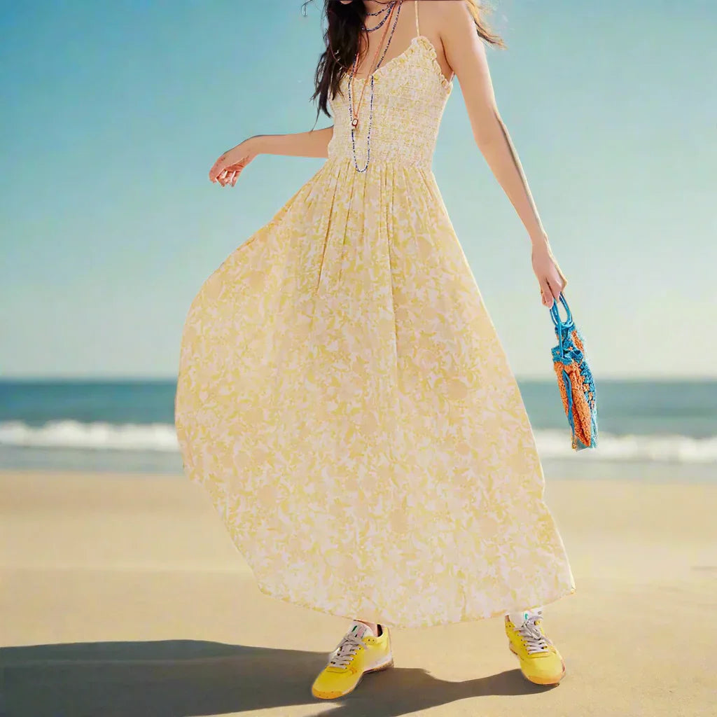 Floral Yellow - A woman wearing a pink and white floral print maxi dress featuring a smocked bodice, ruffled neckline, and thin straps, standing against a rustic stone wall.