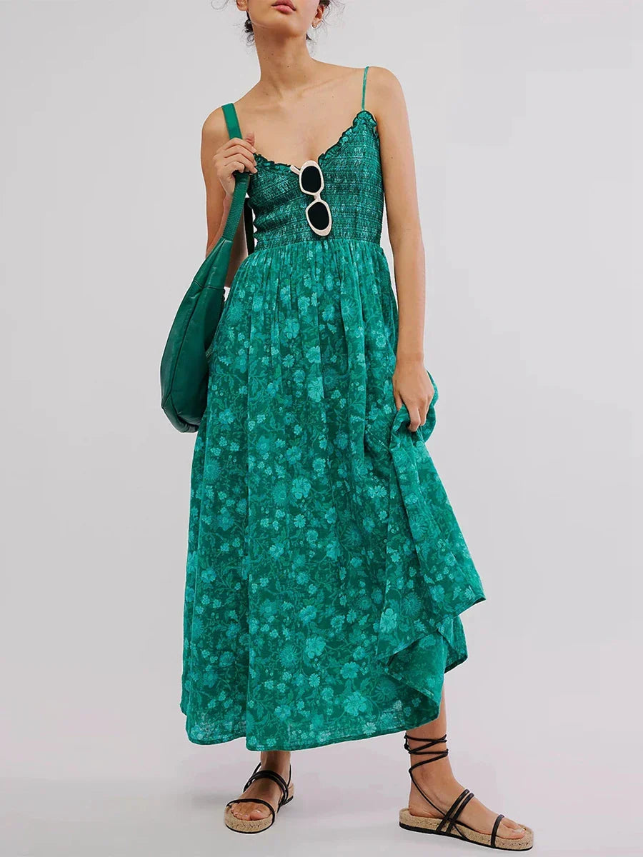 Floral Cyan - A woman wearing a pink and white floral print maxi dress featuring a smocked bodice, ruffled neckline, and thin straps, standing against a rustic stone wall.