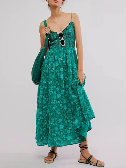 Floral Cyan - A woman wearing a pink and white floral print maxi dress featuring a smocked bodice, ruffled neckline, and thin straps, standing against a rustic stone wall.