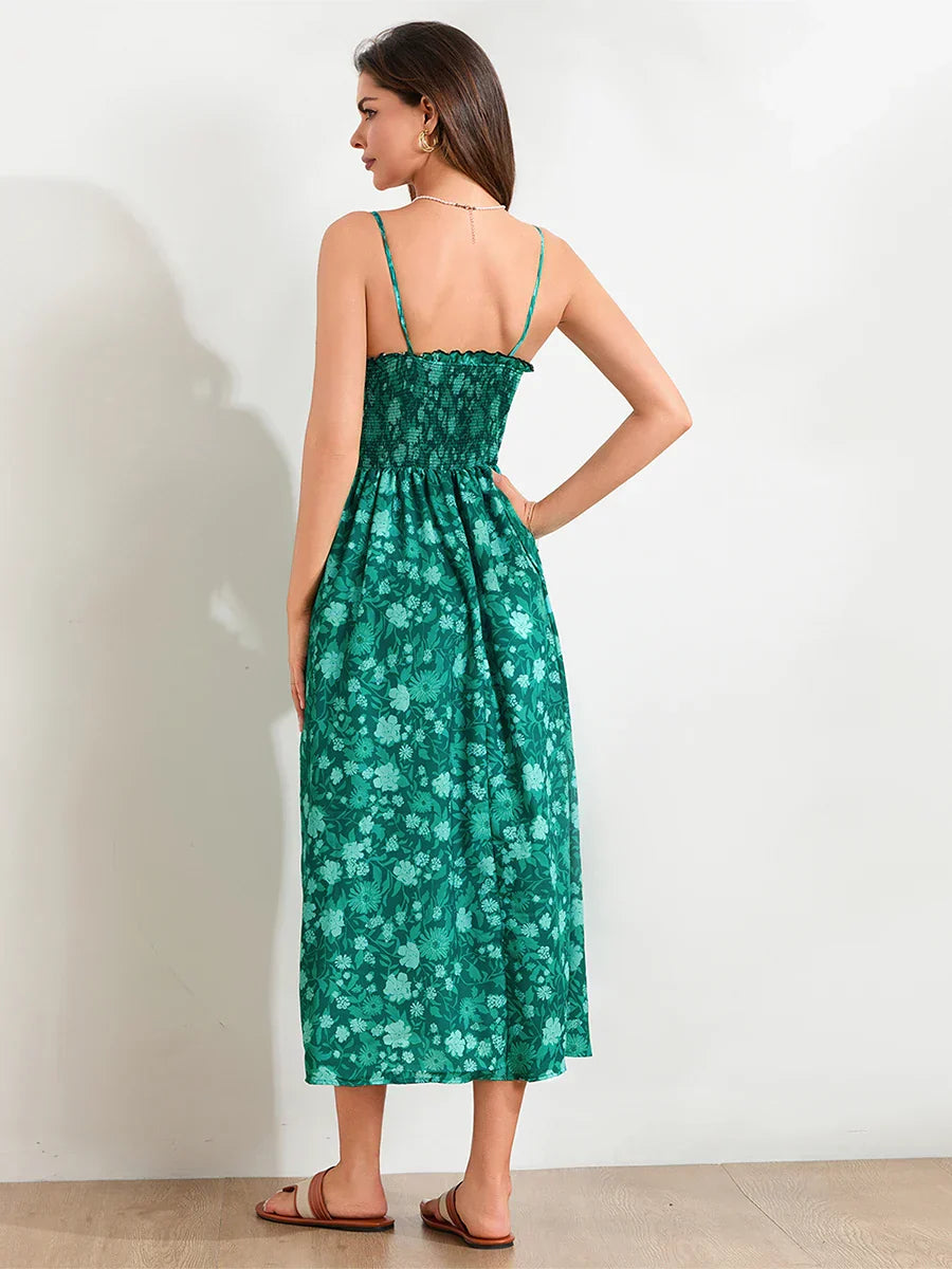 Floral Cyan - A woman wearing a pink and white floral print maxi dress featuring a smocked bodice, ruffled neckline, and thin straps, standing against a rustic stone wall.