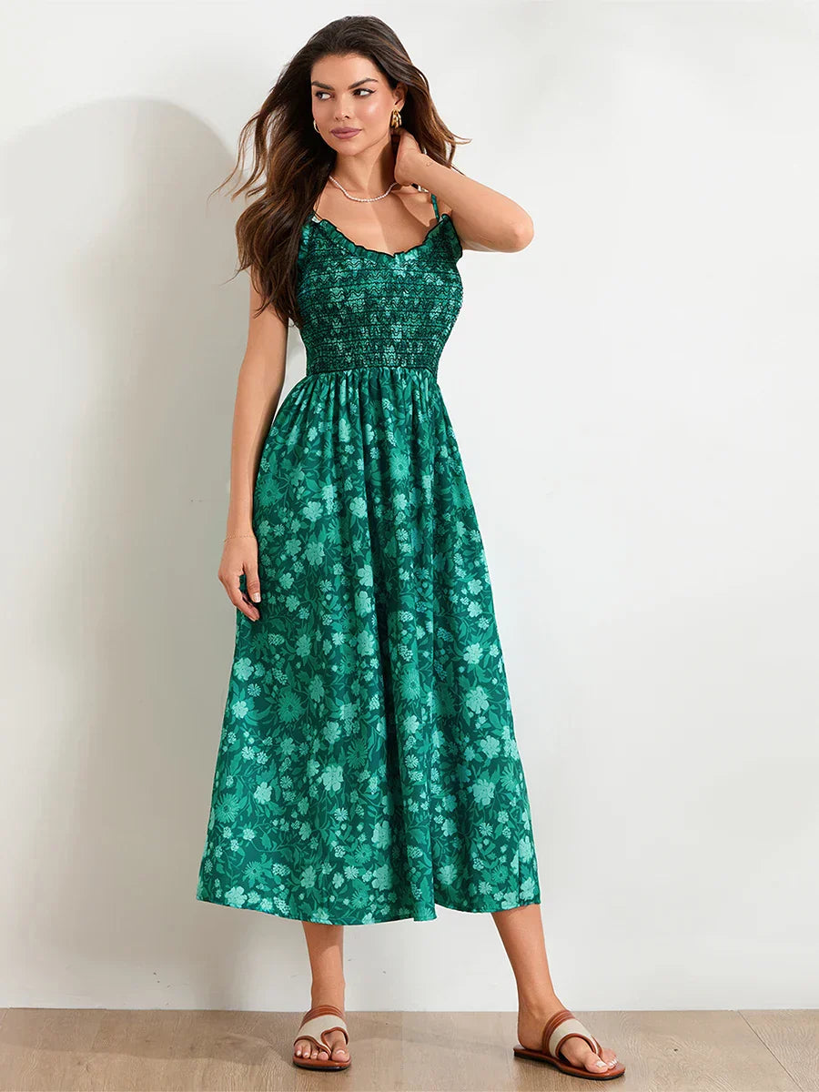 Floral Cyan - A woman wearing a pink and white floral print maxi dress featuring a smocked bodice, ruffled neckline, and thin straps, standing against a rustic stone wall.