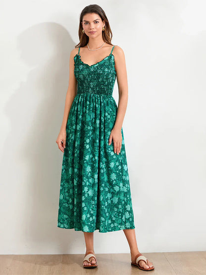 Floral Cyan - A woman wearing a pink and white floral print maxi dress featuring a smocked bodice, ruffled neckline, and thin straps, standing against a rustic stone wall.