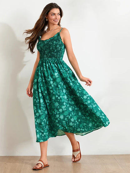 Floral Cyan - A woman wearing a pink and white floral print maxi dress featuring a smocked bodice, ruffled neckline, and thin straps, standing against a rustic stone wall.